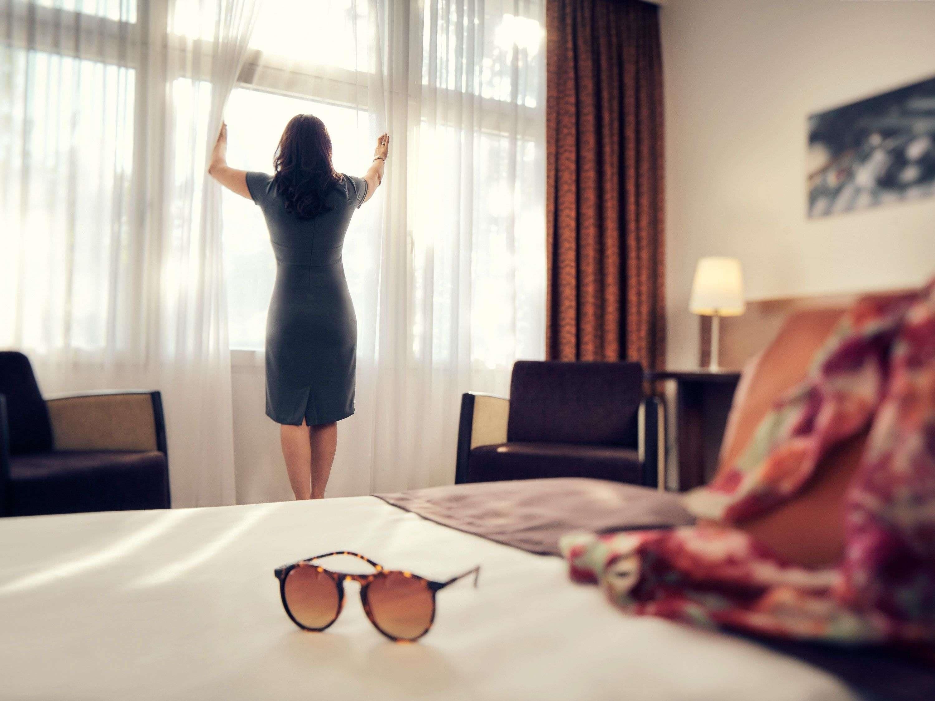 Una habitación con una cama, unos anteojos de sol sobre la cama y una persona de pie junto a una ventana.