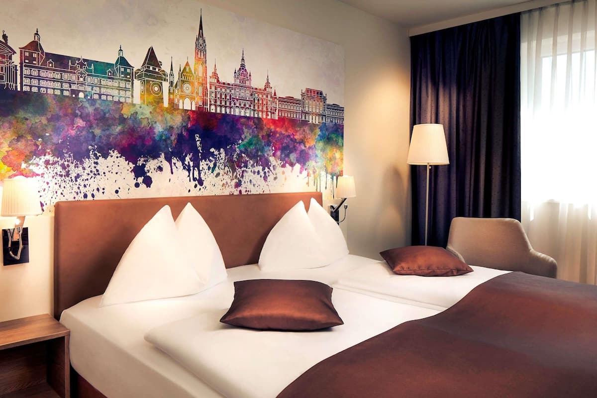 A hotel room with a large bed, a headboard, a nightstand, a lamp, a chair, and a colorful cityscape painting on the wall.