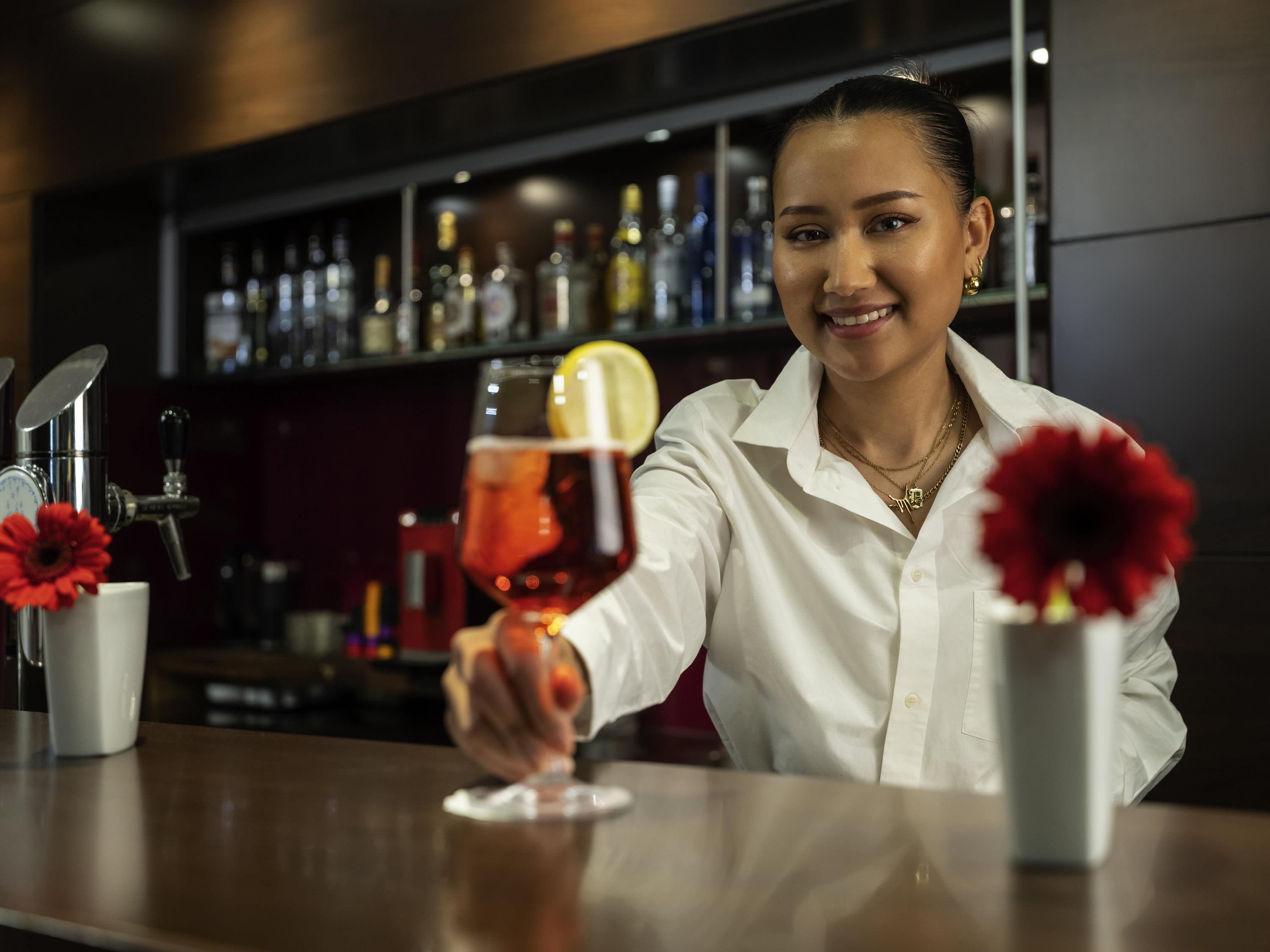Un bar con un cantinero sosteniendo una bebida, una estantería llena de botellas y una decoración floral roja.