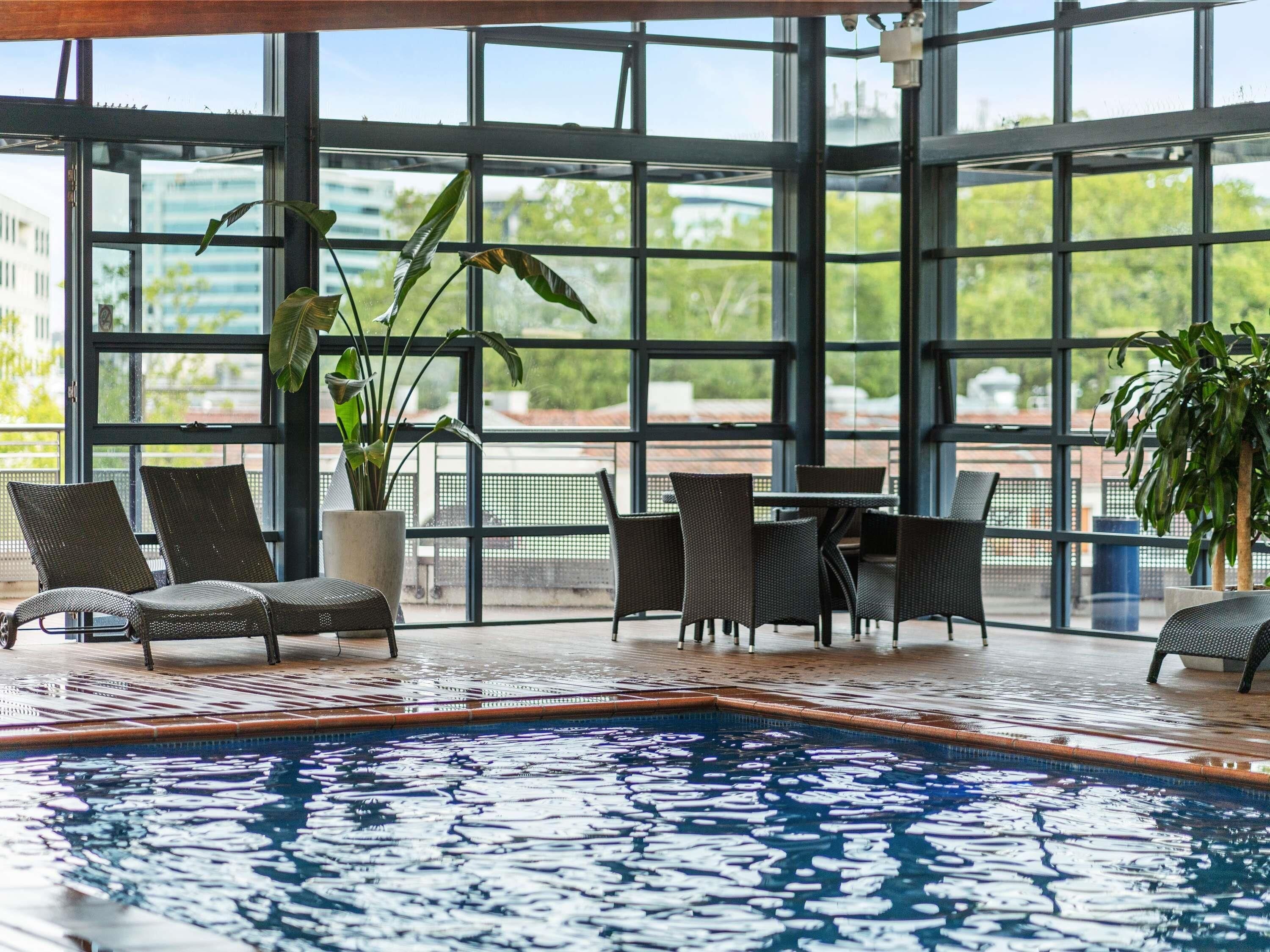 Una piscina interior con paredes de vidrio transparente, mobiliario de mimbre y plantas en macetas.