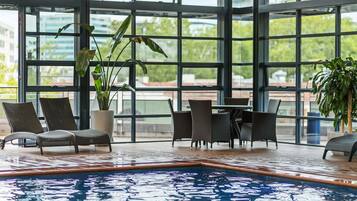 Una piscina interior con paredes de vidrio transparente, mobiliario de mimbre y plantas en macetas.