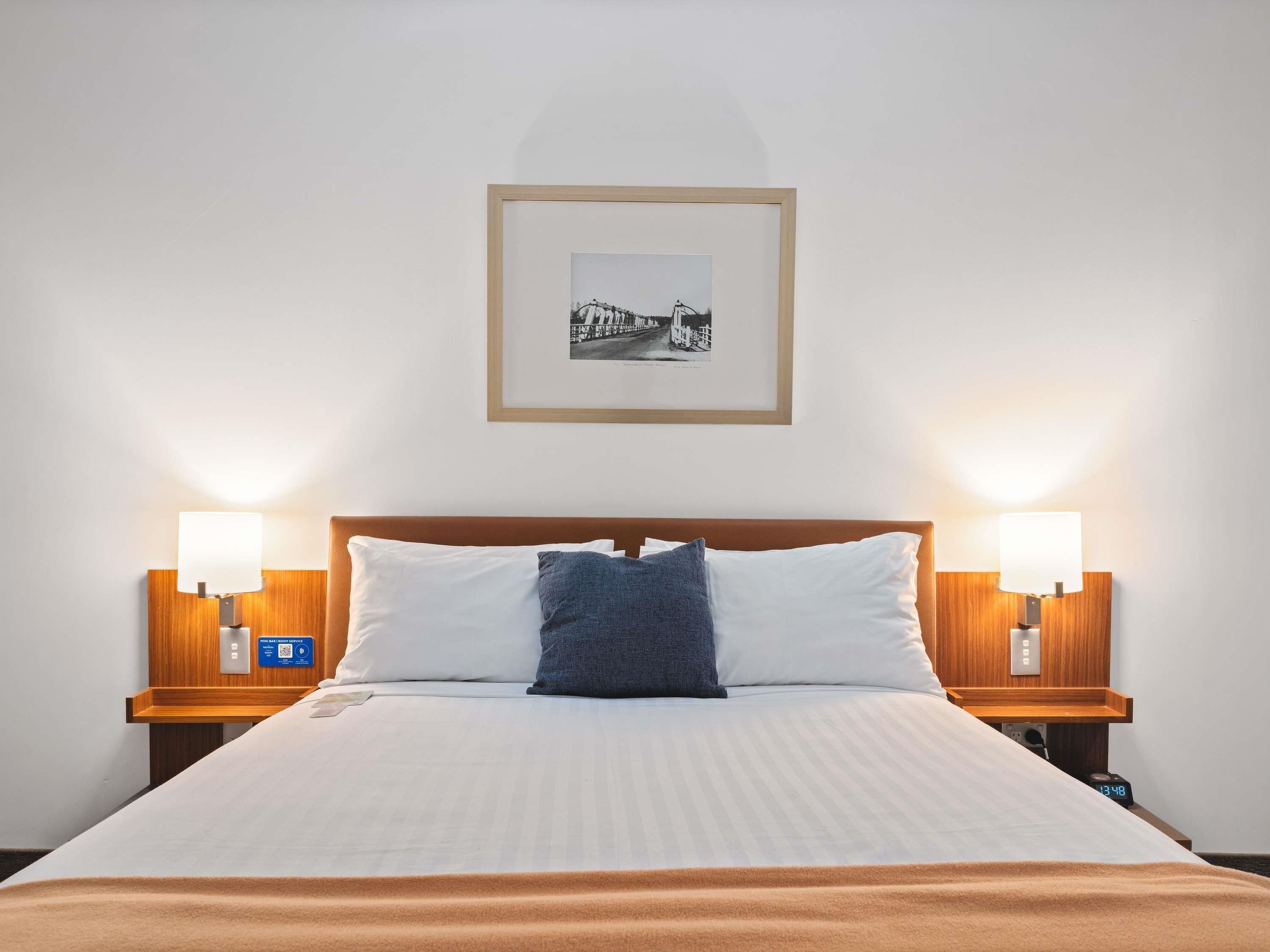 Habitación de hotel con cama, dos lámparas de noche, un cuadro en la pared y un reloj digital en la mesita.
