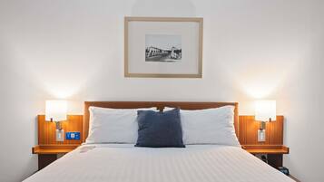 Habitación de hotel con cama, dos lámparas de noche, un cuadro en la pared y un reloj digital en la mesita.