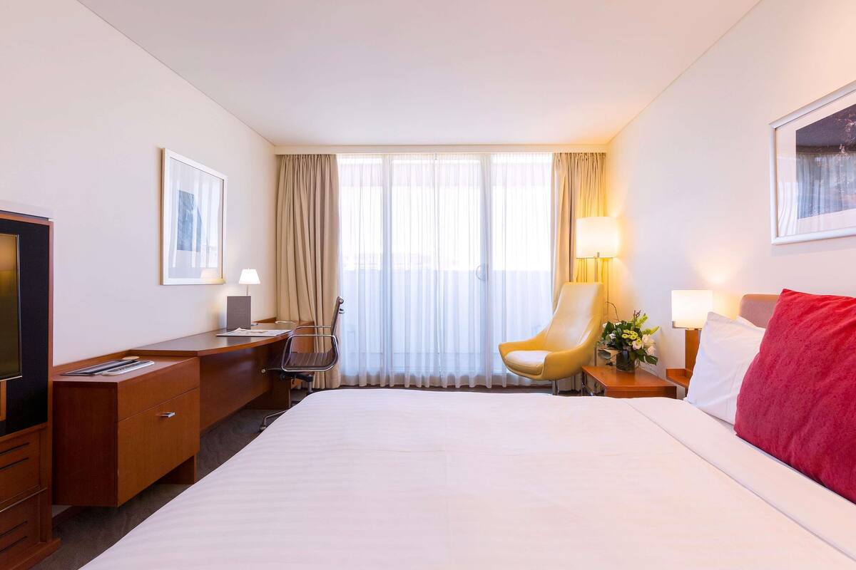 A hotel room with a bed, a desk, a chair, a TV, and a window with curtains.