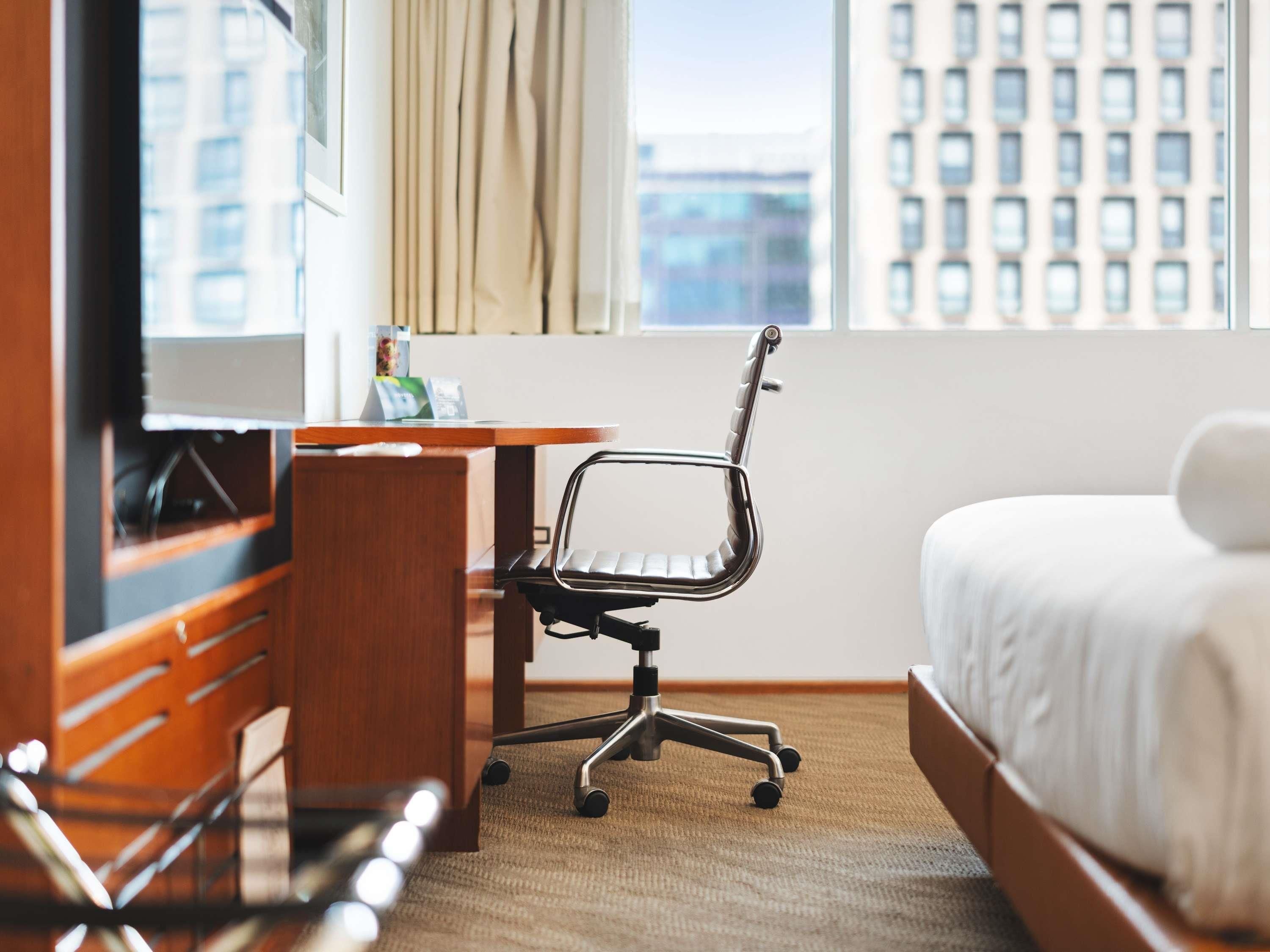 A hotel room with a bed, a desk with a chair, a TV, and a window with curtains.