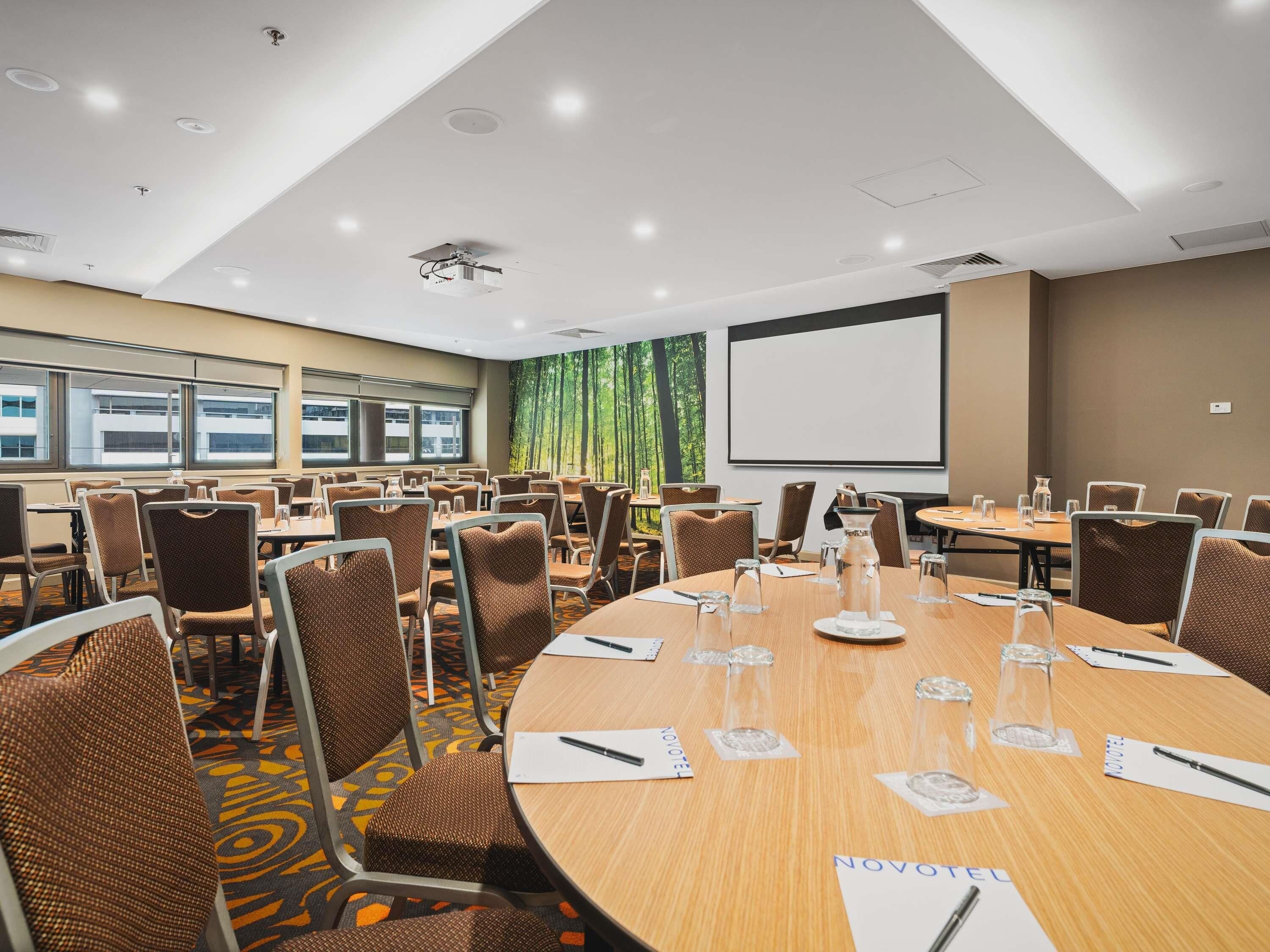 A conference room with round tables, brown chairs, a projector screen, and a large window with a view of trees.