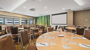 A conference room with round tables, brown chairs, a projector screen, and a large window with a view of trees.