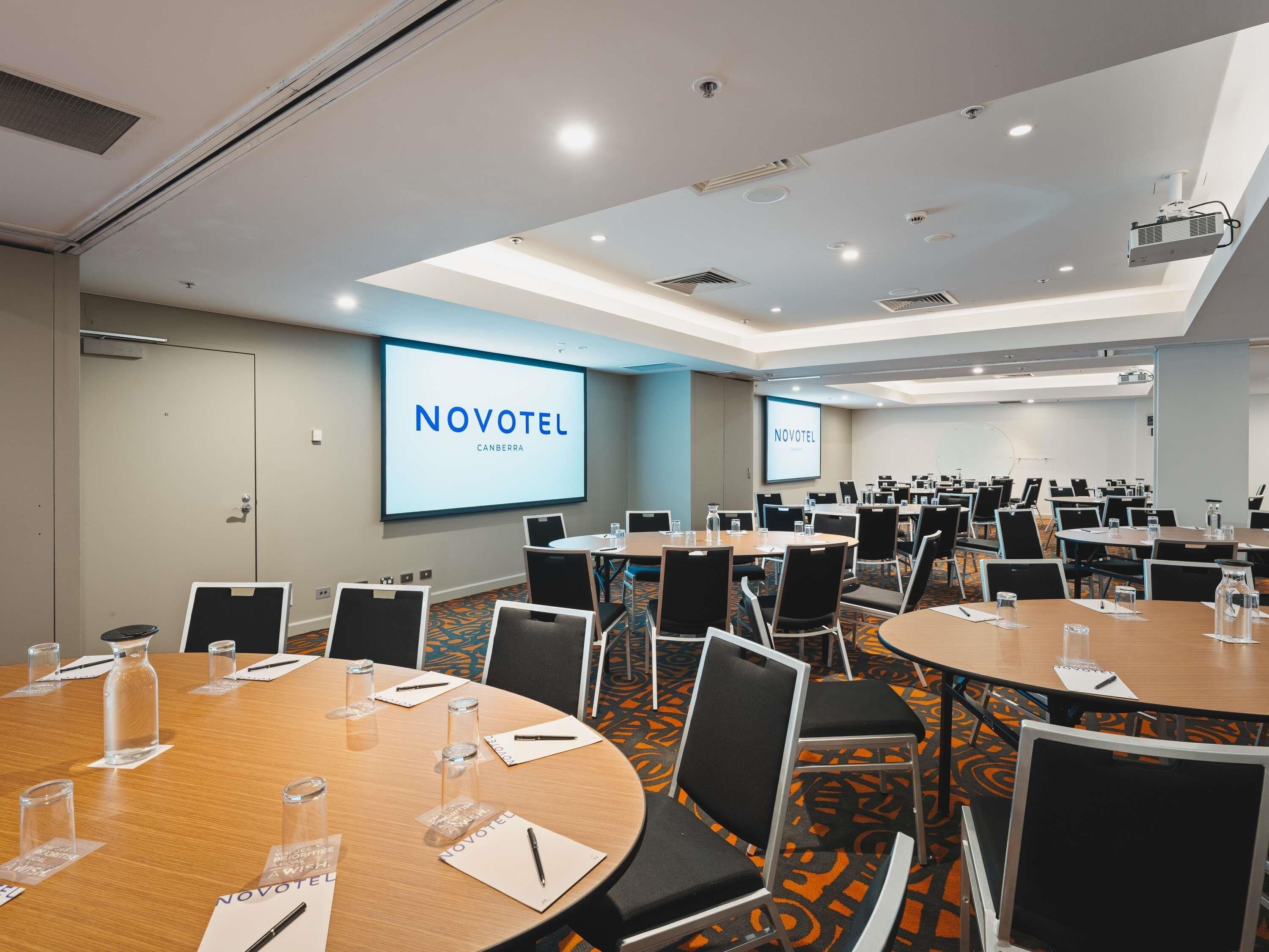 A conference room with multiple tables, chairs, and a large screen displaying 'NOVOTEL CANBERRA'.