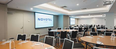 A conference room with multiple tables, chairs, and a large screen displaying 'NOVOTEL CANBERRA'.