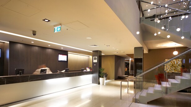 A modern hotel reception area with a marble counter, a digital screen, and a staircase leading to an upper level. - Novotel Canberra (Canberra)