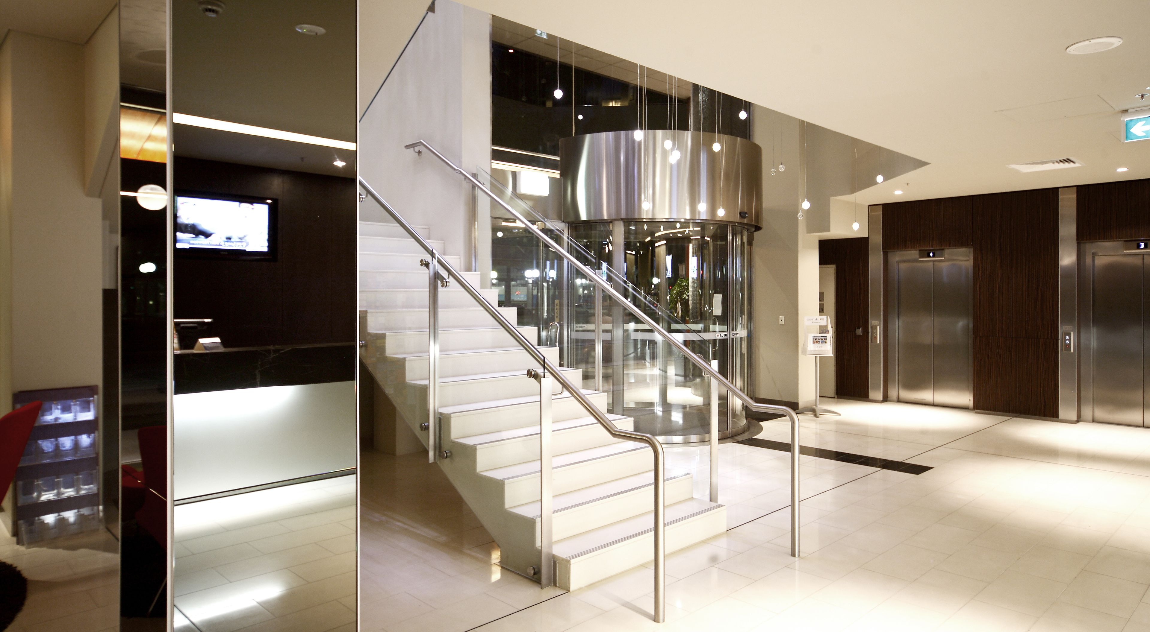 A modern hotel lobby with a glass staircase, a reception desk, and a large cylindrical light fixture.