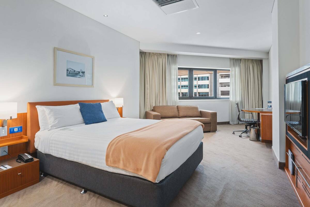A hotel room with a bed, a desk, a chair, a TV, and a window with curtains.