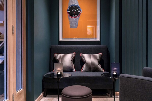 A modern lounge area with a sofa, two pillows, a small table with a candle, and a framed watch on the wall.