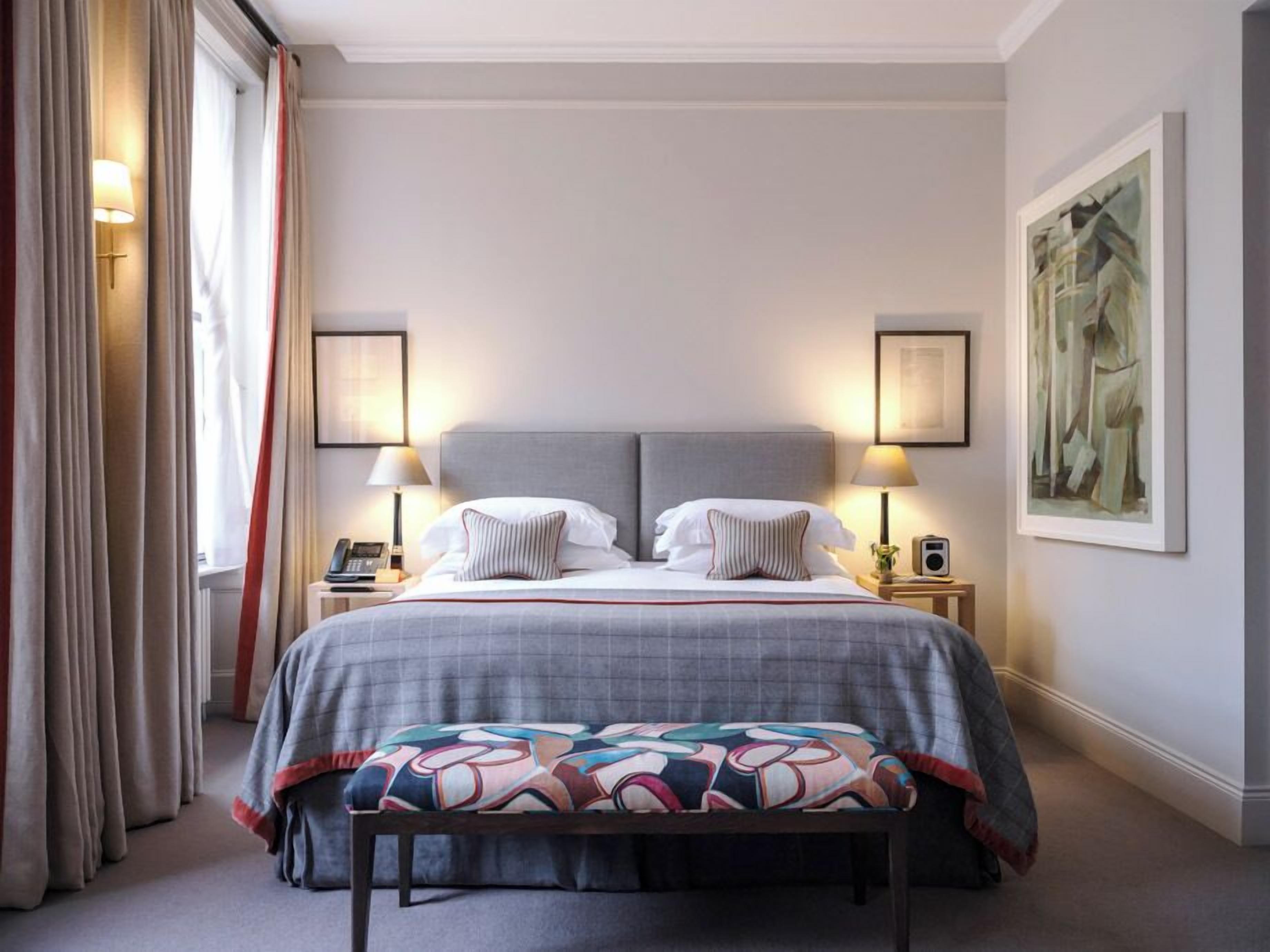 A hotel room with a large bed, two bedside lamps, a bedside telephone, a bench, and a painting on the wall.