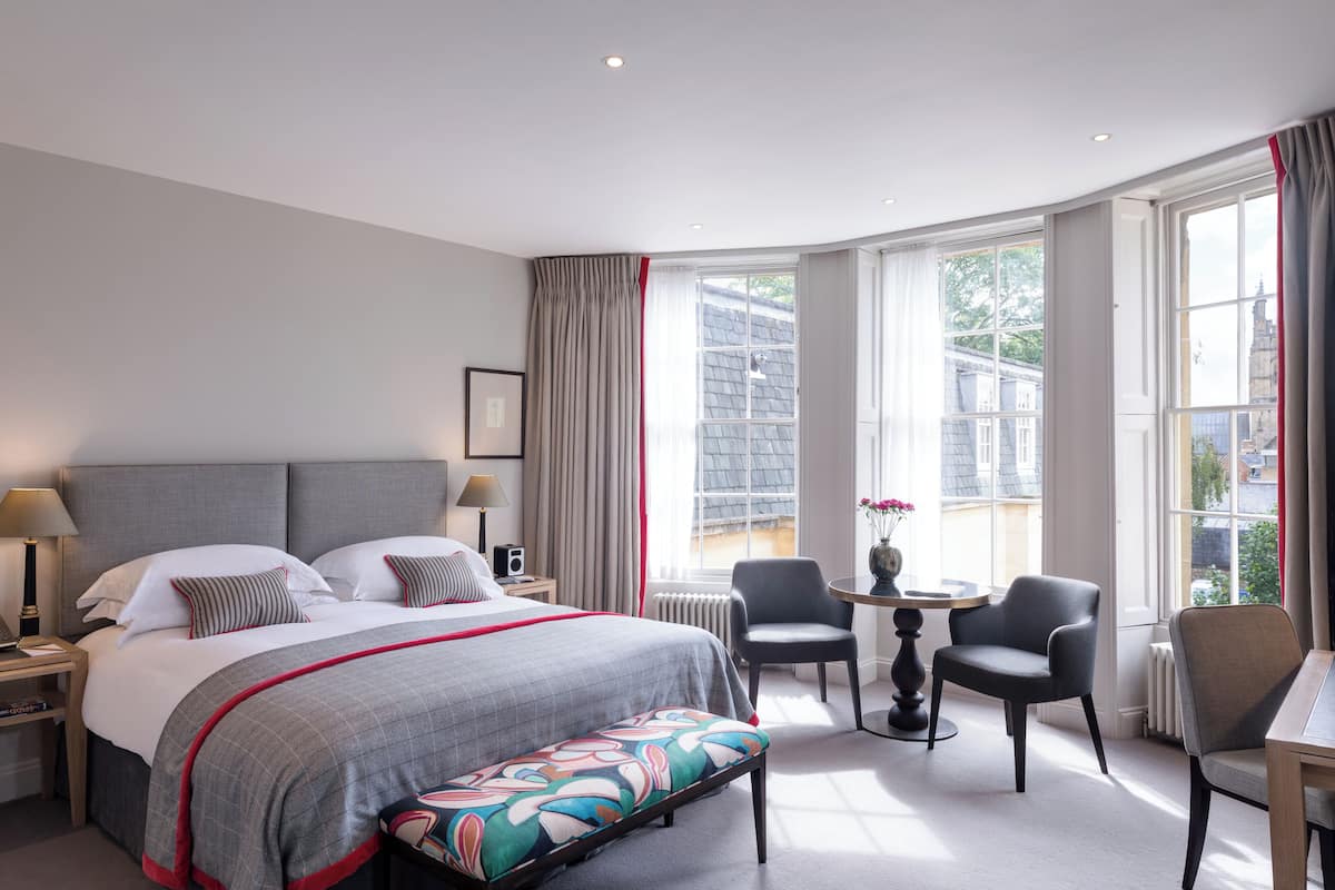 A hotel room with a large bed, a desk, two chairs, and a view of the city through large windows.