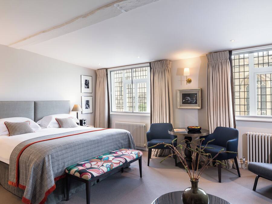 Une chambre d’hôtel comprenant un lit, un bureau, deux chaises, un banc et un vase avec des branches.