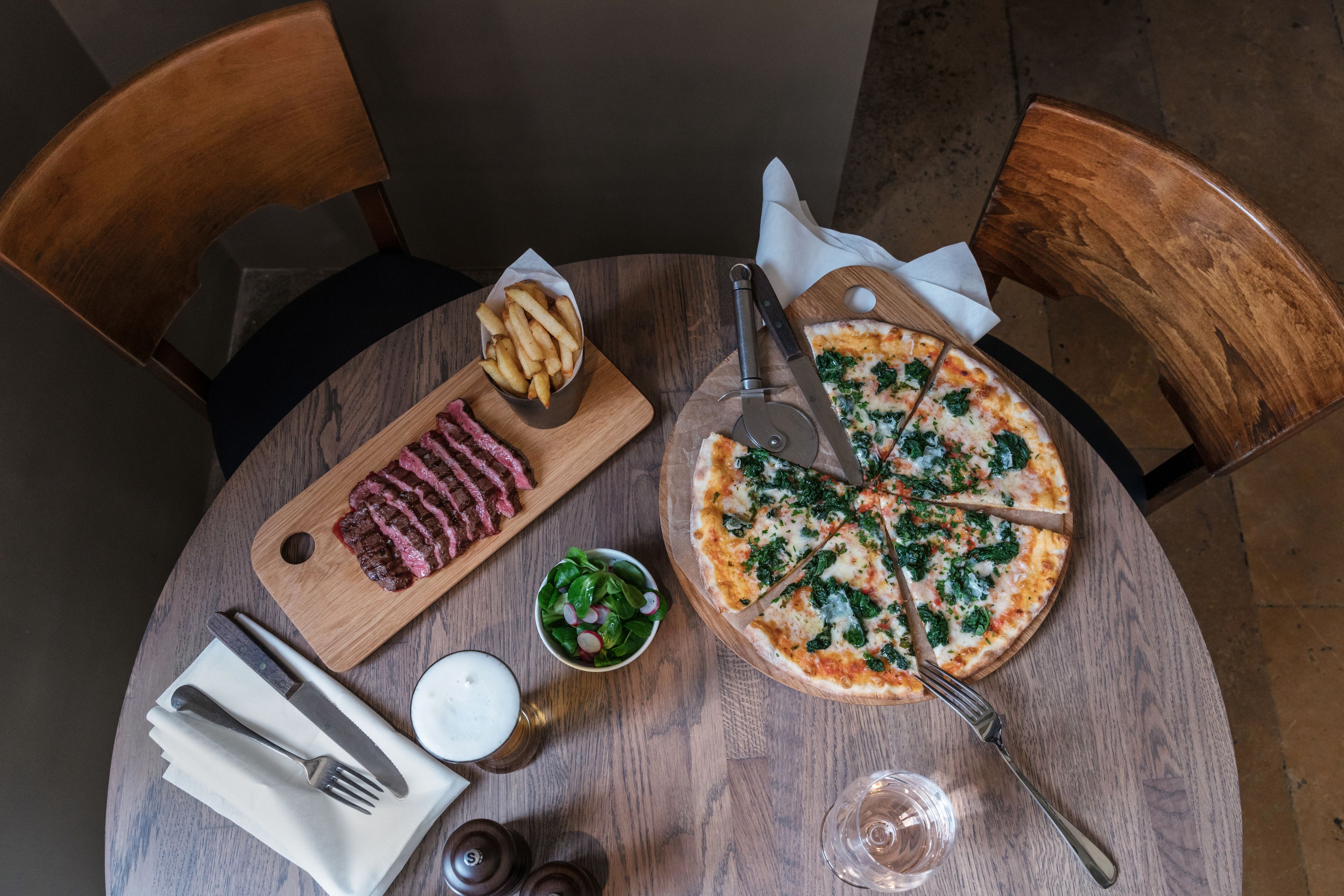Puisesta pöydästä löytyy pizza, kulho salaattia, lautanen pihviä ja lasi olutta.