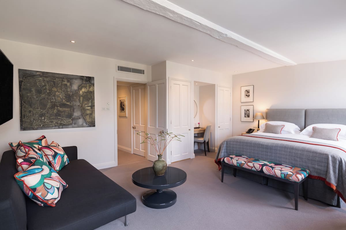 A modern hotel room with a large bed, a sofa, a small round table with a vase, and a desk with a chair.