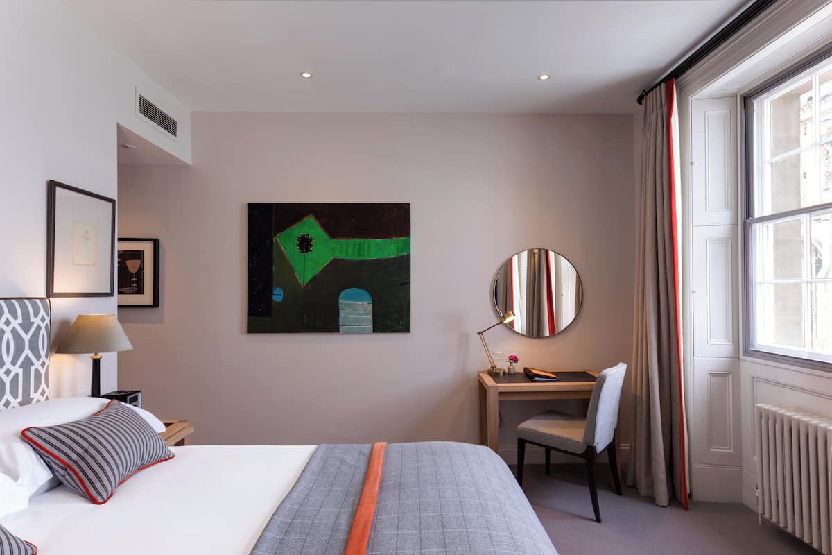 A hotel room with a bed, a desk, a chair, a large window with curtains, and a painting on the wall.