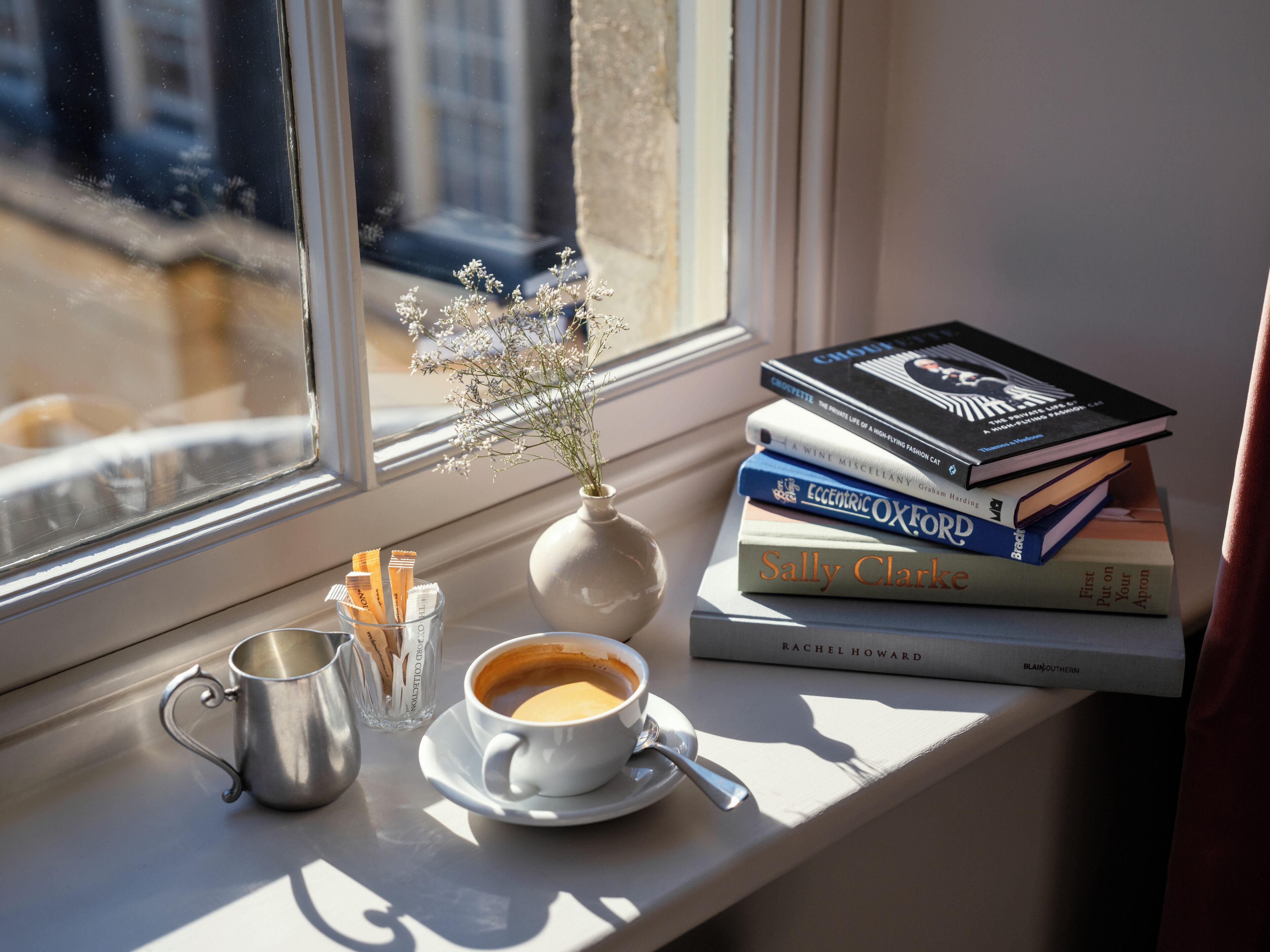 A window sill with a cup of coffee, a vase with flowers, a stack of books, and a metal mug.
