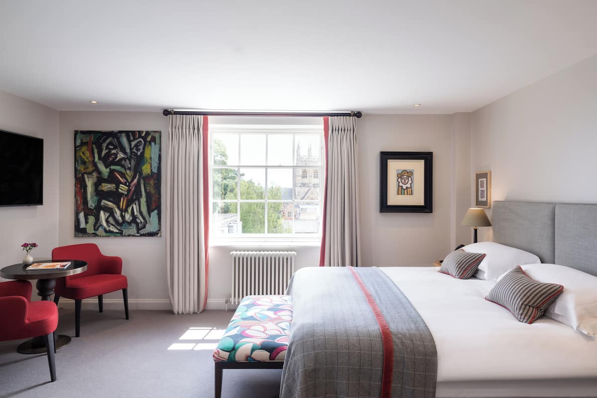 A hotel room with a large bed, a desk, a chair, a radiator, and a window with curtains.