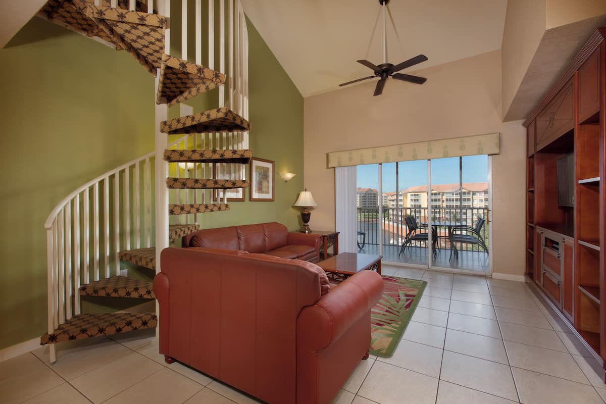 A living room with a brown leather sofa, a staircase with patterned railings, a ceiling fan, and a view of a balcony with outdoor furniture.