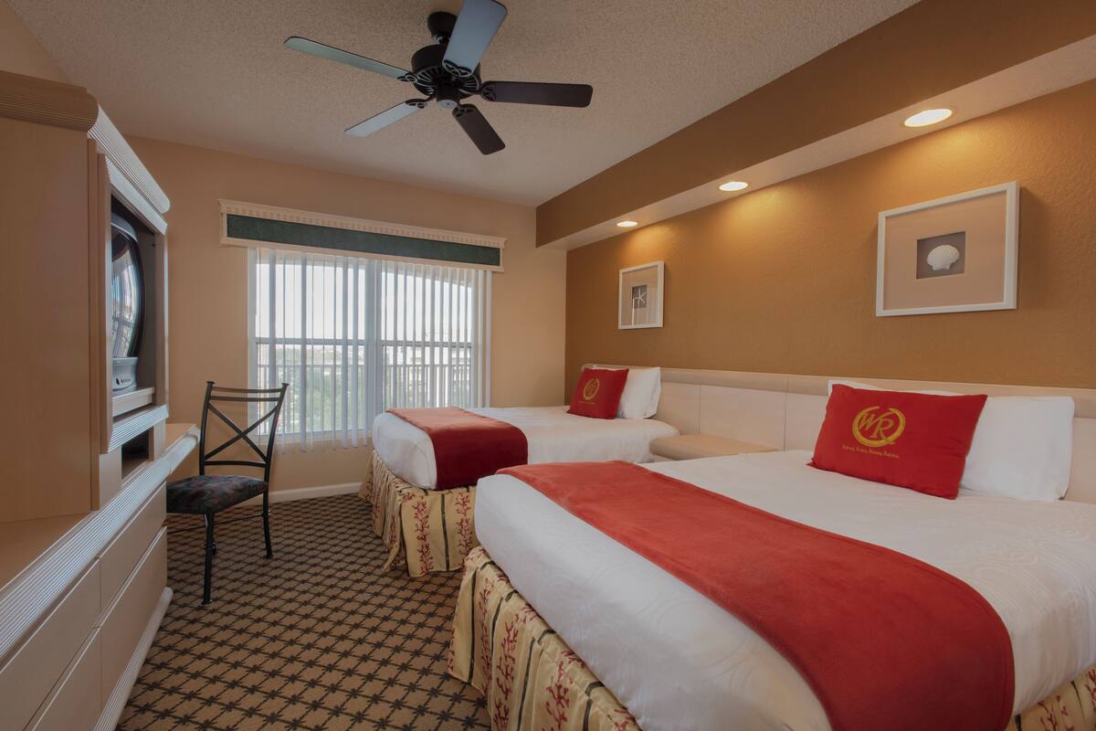 A hotel room with two beds, a ceiling fan, a television, and a window with blinds.