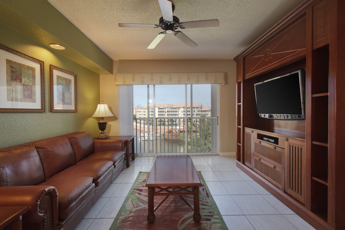 A living room with a brown leather sofa, a wooden coffee table, a ceiling fan, a TV on a wooden cabinet, and a balcony with a view of buildings.