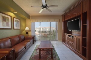 A living room with a brown leather sofa, a wooden coffee table, a ceiling fan, a TV on a wooden cabinet, and a balcony with a view of buildings.