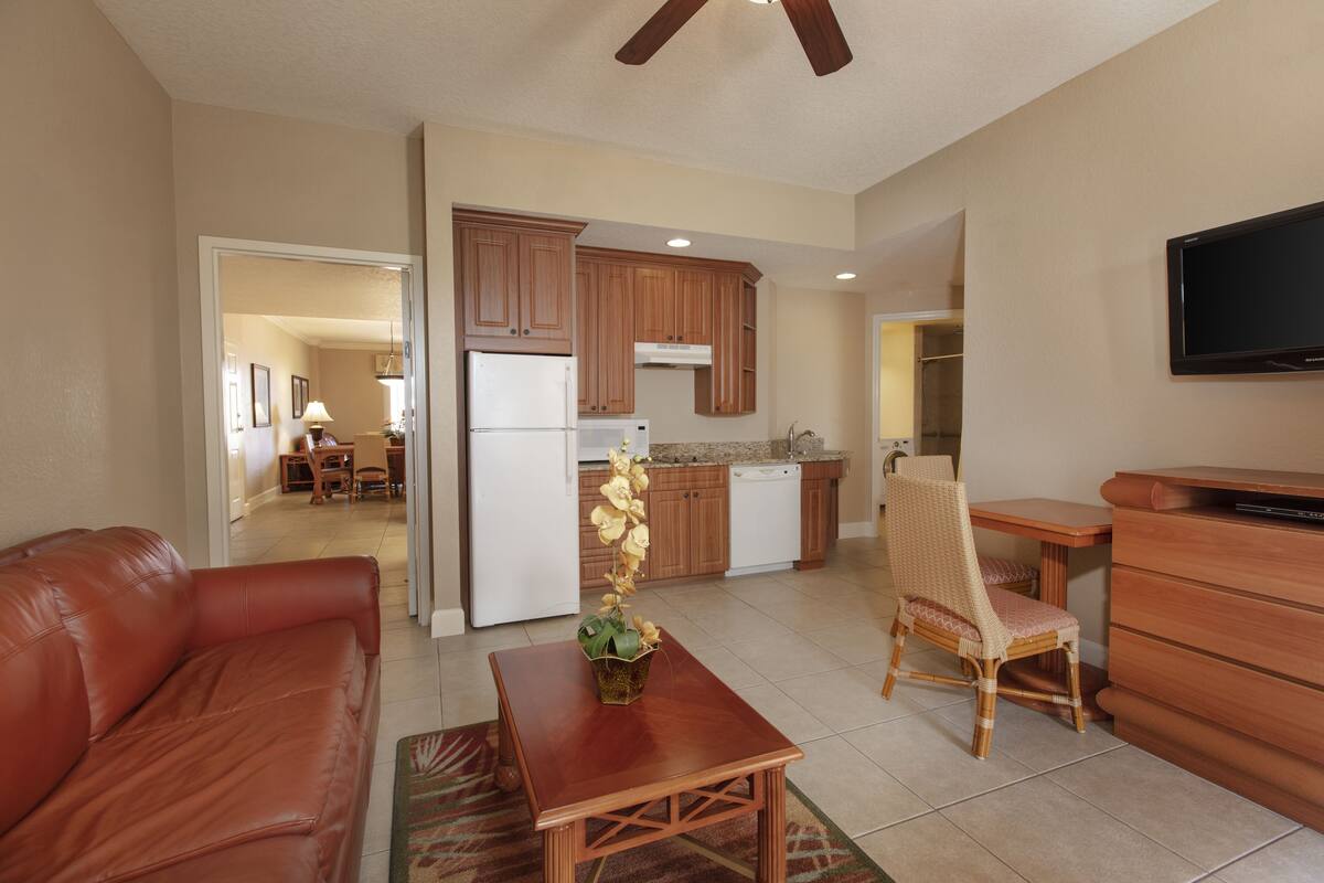 A living room with a brown leather sofa, a wooden coffee table, and a wicker chair. There is a kitchen area with wooden cabinets, a refrigerator, and a dishwasher. A flat-screen TV is mounted on the wall.