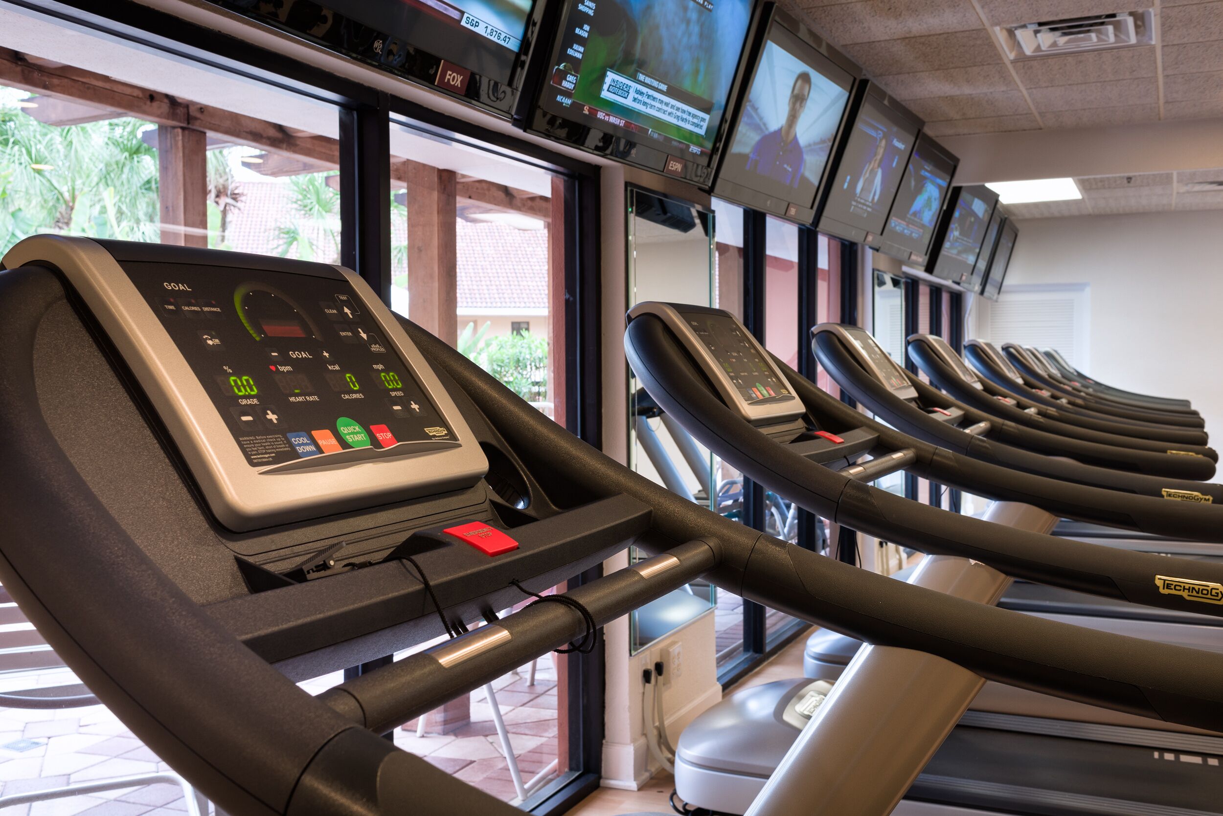 File di tapis roulant con display digitali, una finestra con vista su palme e diversi schermi sopra i tapis roulant.