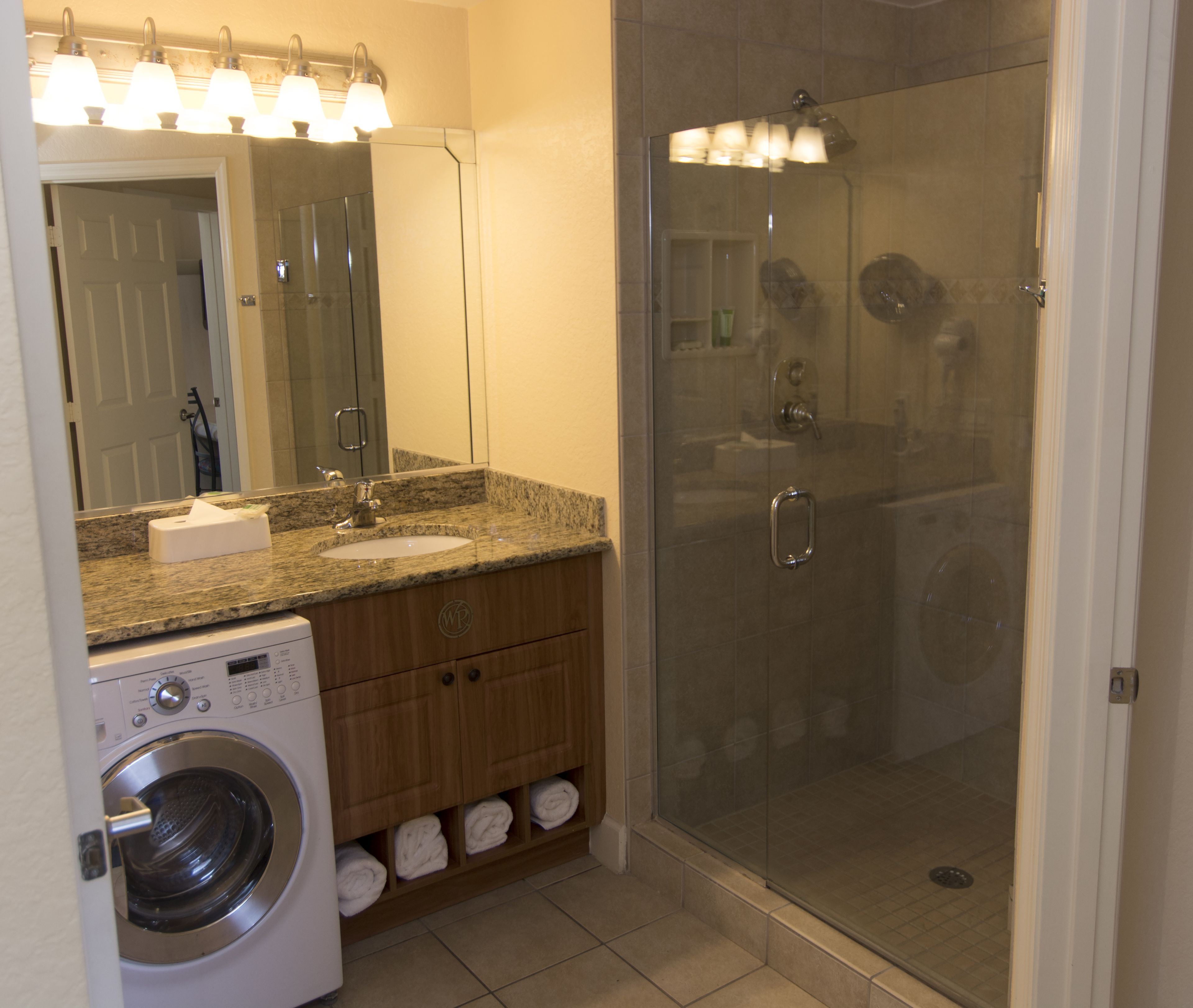 A modern bathroom with a granite countertop, a built-in washing machine, and a glass-enclosed shower.