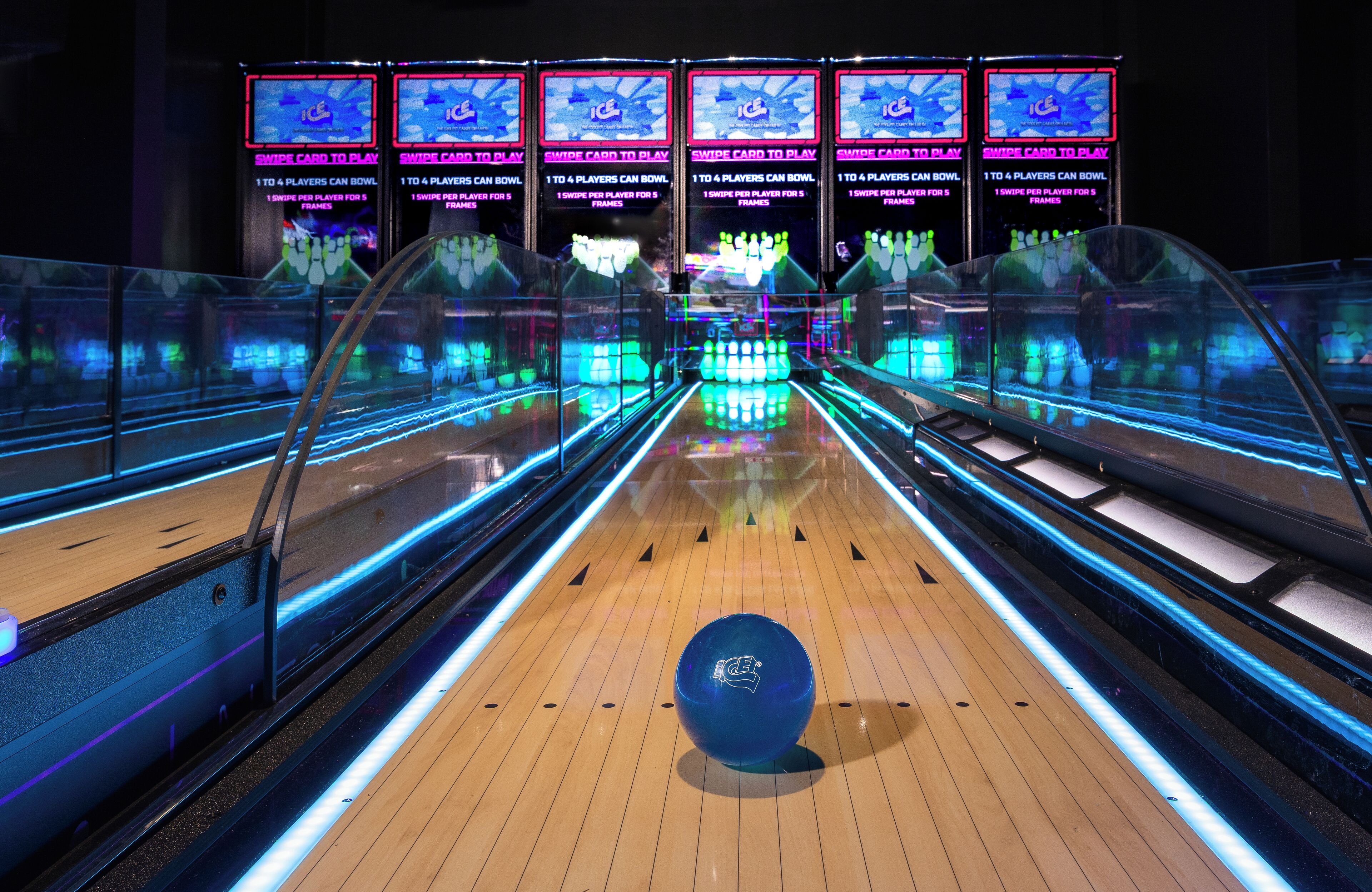 A bowling alley with multiple lanes, illuminated by blue and green lights, and a bowling ball in the center of the frame.