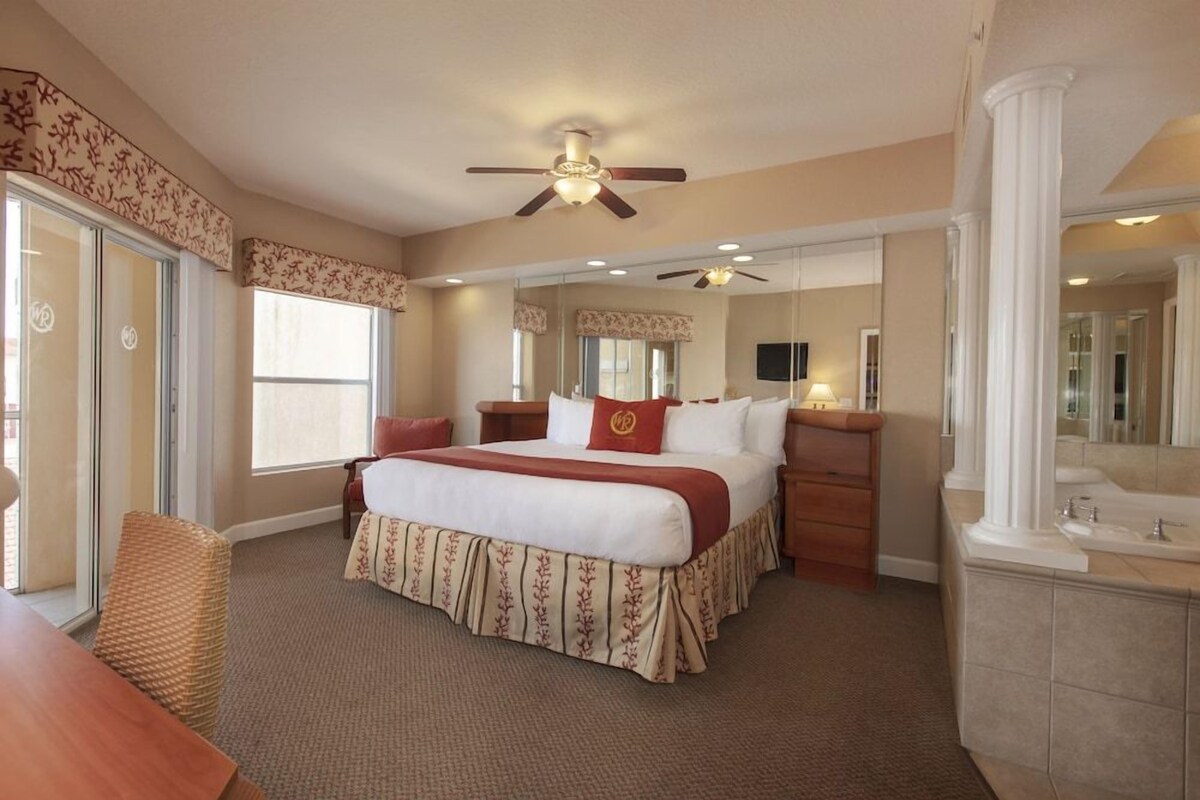 Une chambre d’hôtel avec un grand lit, un bureau, une chaise, un ventilateur de plafond, un miroir et une salle de bain avec un lavabo et un miroir.
