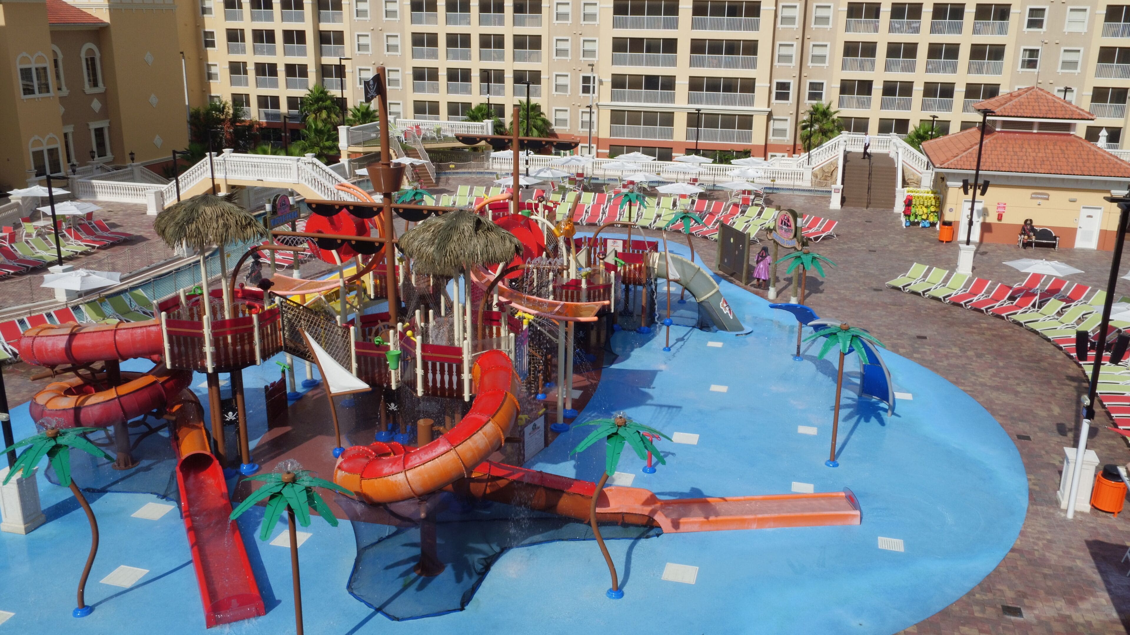 A water park with slides and play structures, surrounded by lounge chairs and umbrellas, with a hotel building in the background.