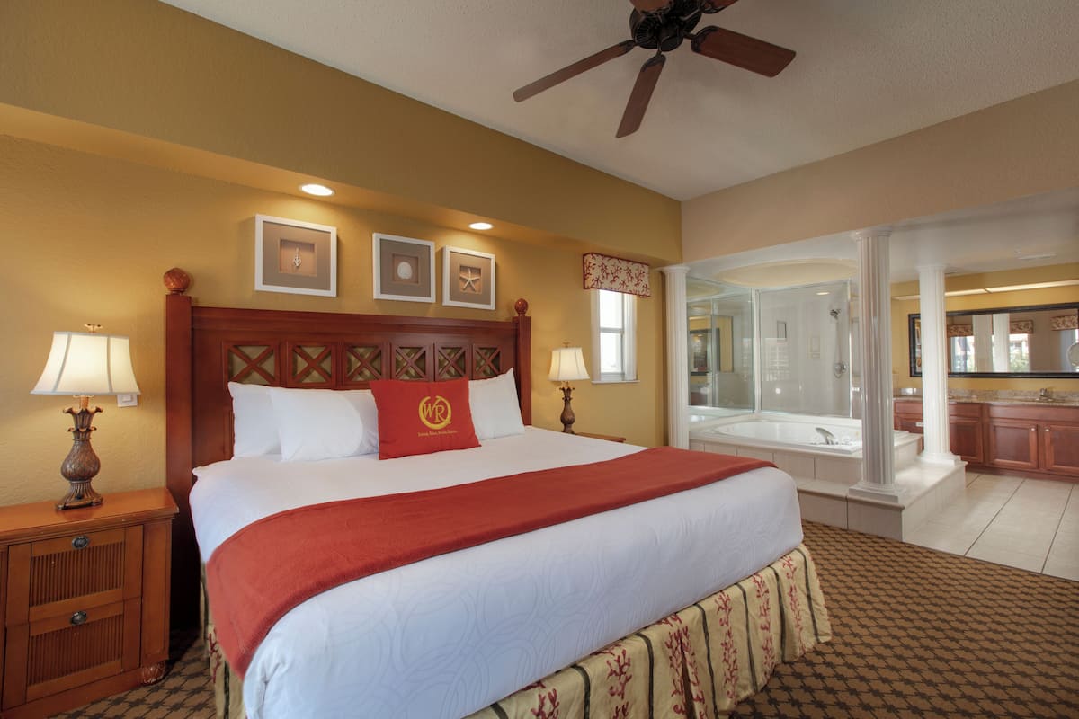 A hotel room with a large bed, two bedside lamps, a ceiling fan, and a bathroom visible through an open door.