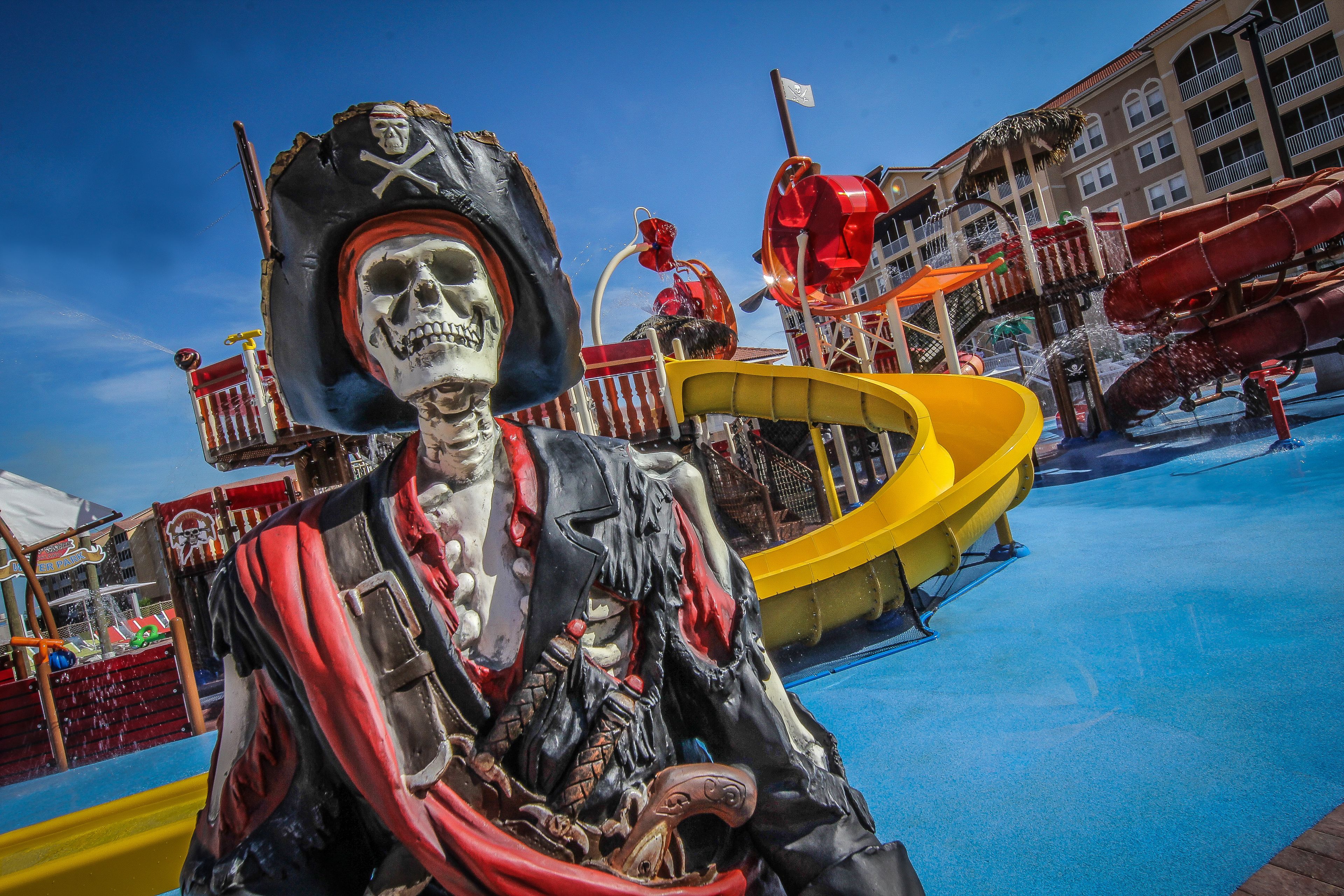 Una figura de pirata esquelético en un parque acuático con toboganes y un edificio al fondo.