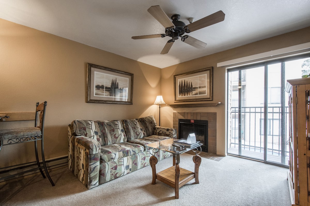 A living room with a patterned sofa, a glass coffee table, a fireplace, and a large window.