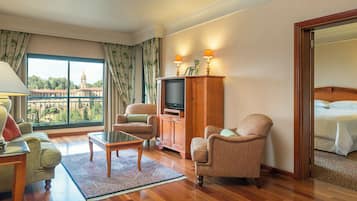 A hotel room with a large window offering a view of a historic building, a television, two armchairs, a small table, and a bed.