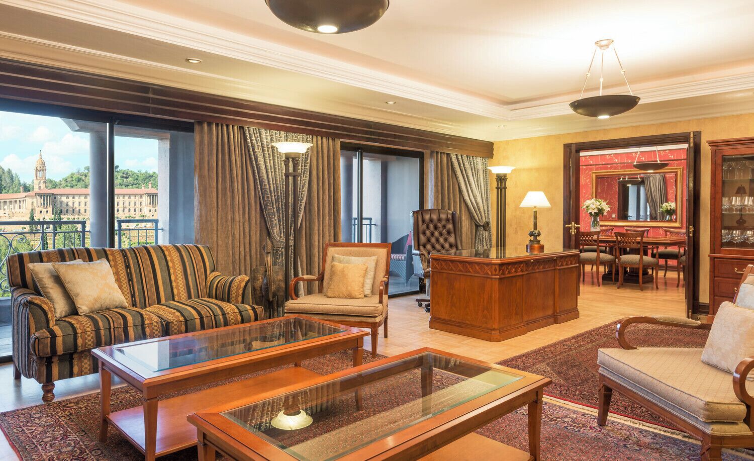 Amplia sala de estar con sofá, sillón y mesa de centro, un escritorio de madera con sillas, y vista a un edificio a través de amplios ventanales.
