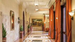 Un pasillo de hotel con piso de mármol, plantas en macetas, un cuadro en la pared y cortinas naranjas.
