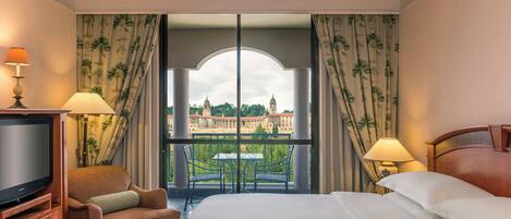 Habitación de hotel con una cama grande, televisión, una silla y un balcón con vistas a un edificio.