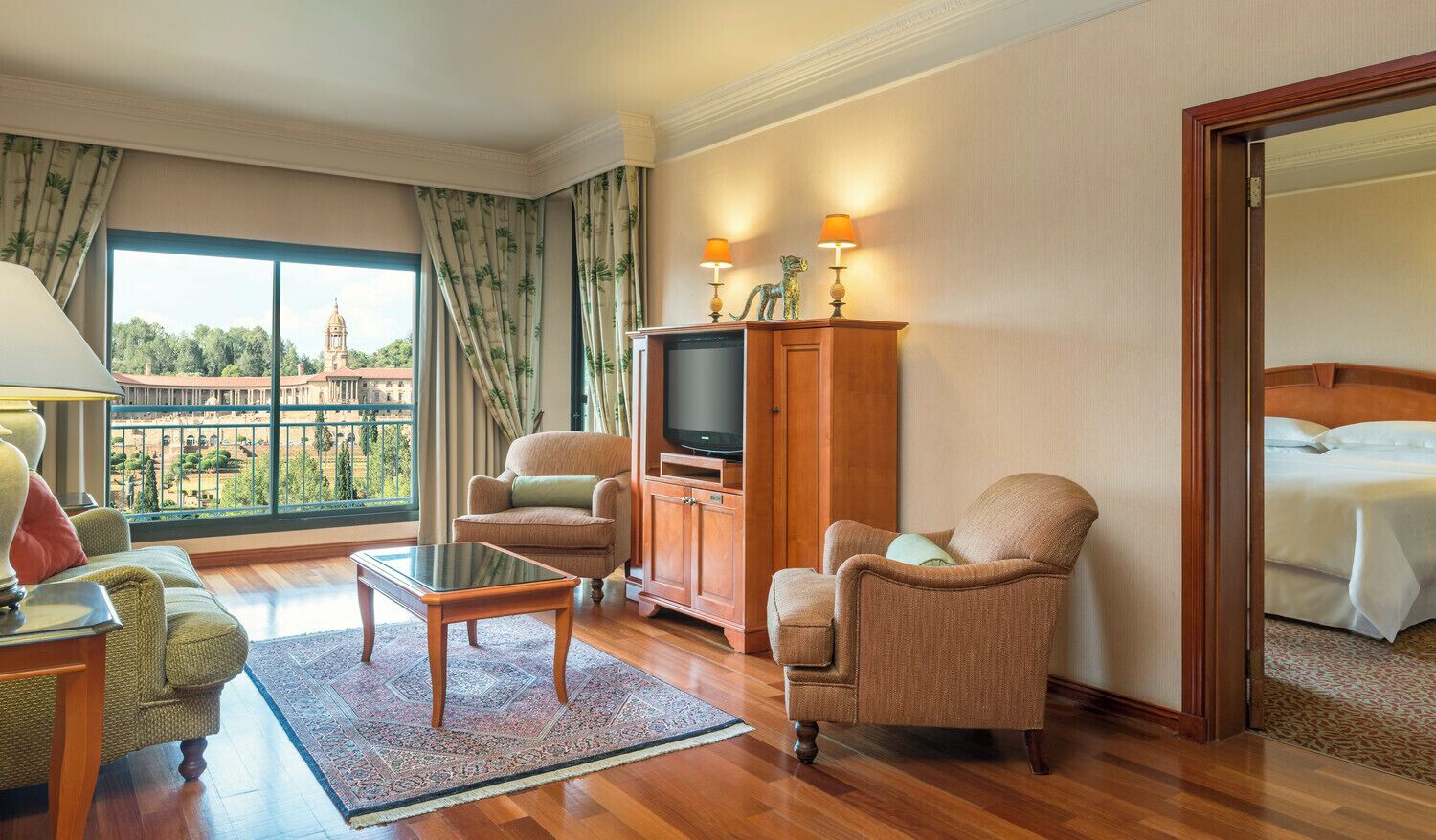 Habitación de hotel con un ventanal grande, televisor, dos sillones, un sofá, un armario de madera y una cama.