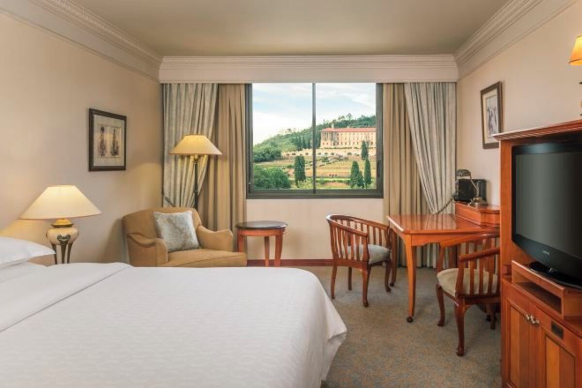 Habitación de hotel con cama, escritorio, silla, televisión y ventana con vistas a un edificio y árboles.