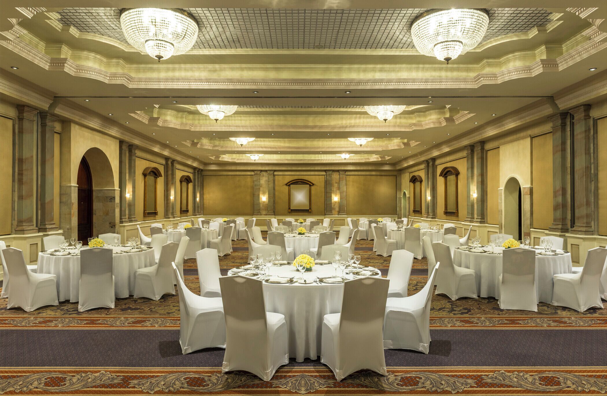 Une salle de banquet avec des tables rondes, des chaises blanches, des lustres et un sol recouvert de moquette.
