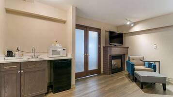 A modern kitchen with a microwave, coffee maker, and a refrigerator. There is a fireplace with a TV mounted above it. A blue armchair and a small table are placed near the fireplace.
