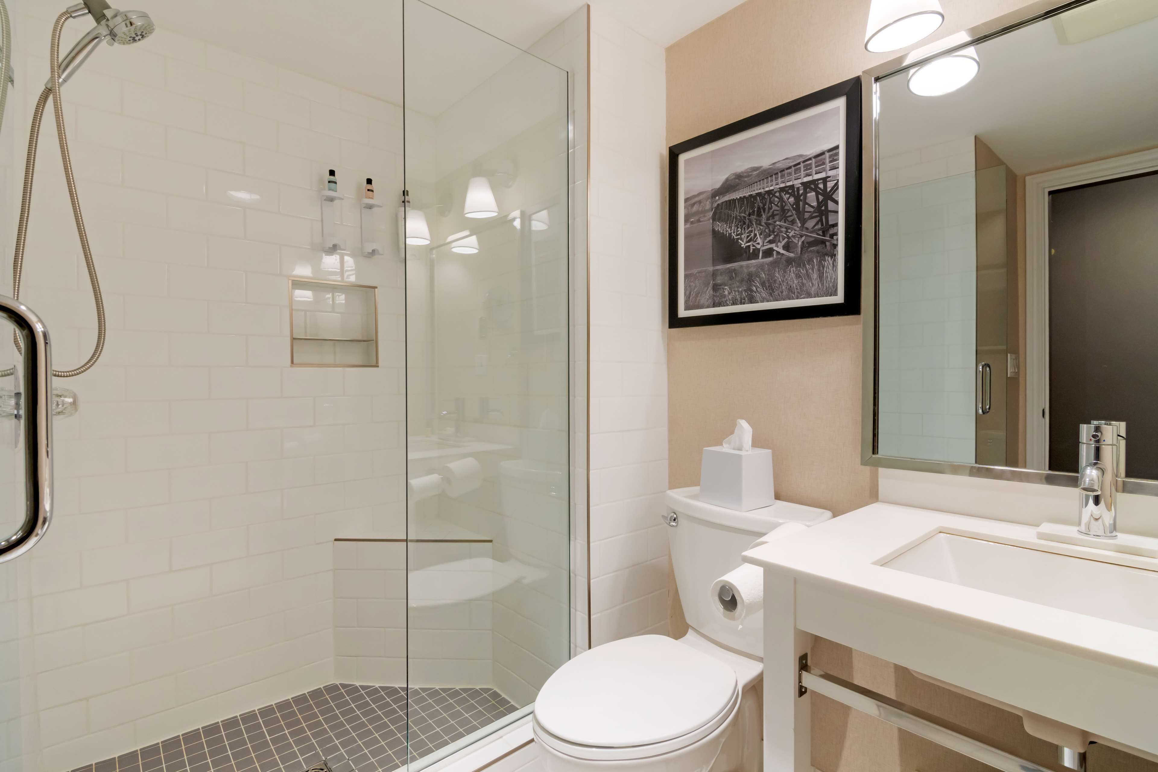 A modern bathroom with a glass-enclosed shower, a white toilet, a sink with a mirror, and a framed picture on the wall.