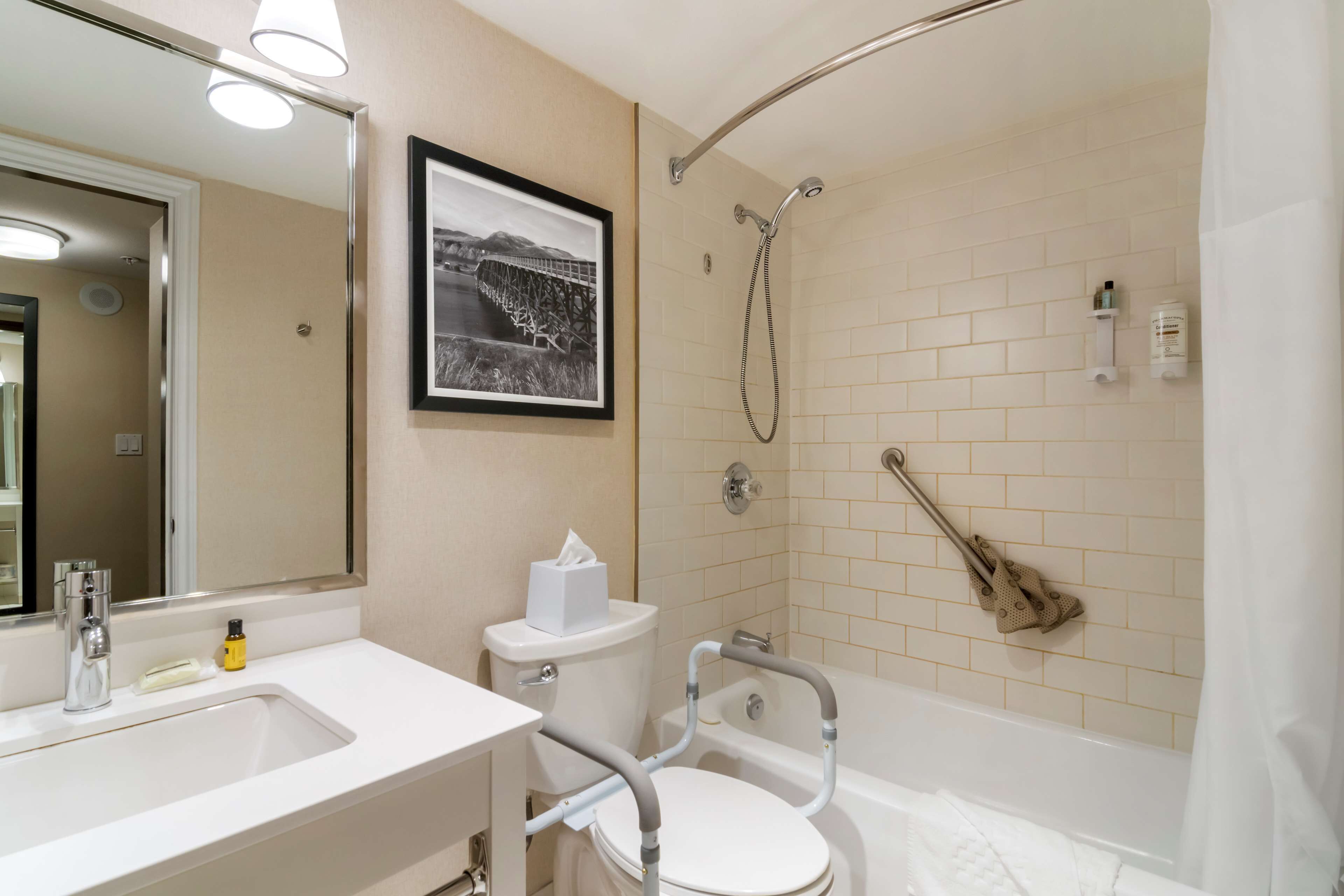 A bathroom with a white sink, toilet, and bathtub. There is a framed picture on the wall, a towel rack, and a handrail for the toilet.