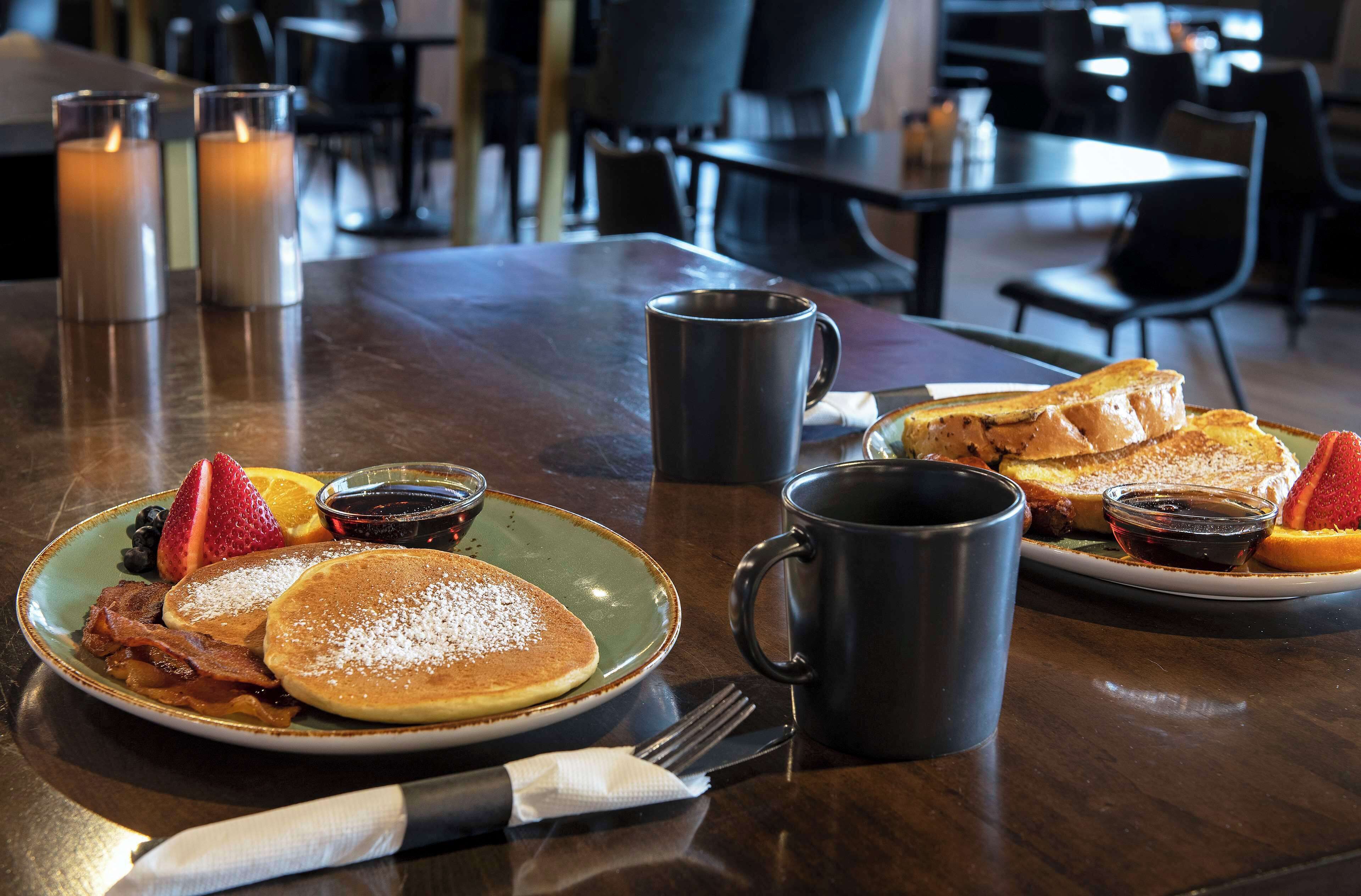 A breakfast setting with pancakes, bacon, strawberries, blueberries, and syrup on a plate, accompanied by a mug and a fork on a wooden table.