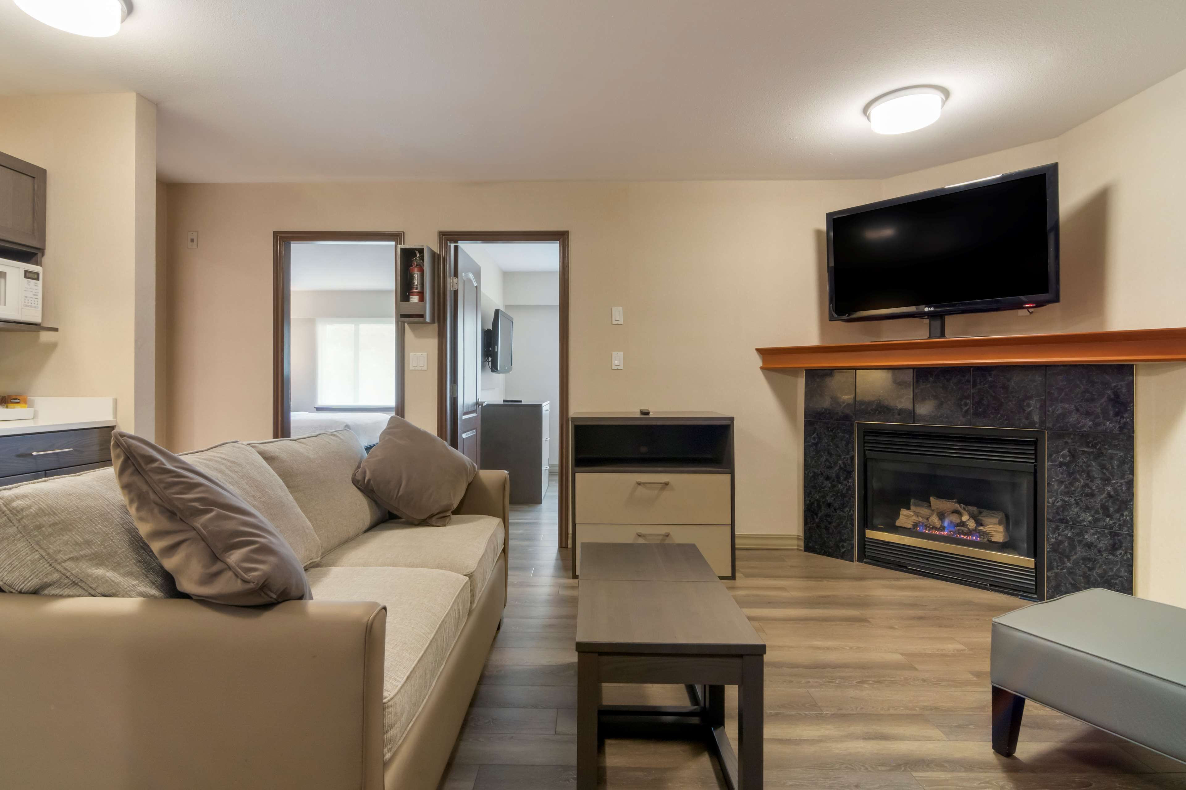 A living room with a sofa, a coffee table, a flat-screen TV mounted above a fireplace, and a kitchen area in the background.